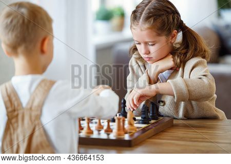 Focused Children Playing Chess Game At Home While Sitting In Living Room At Table With Chessboard