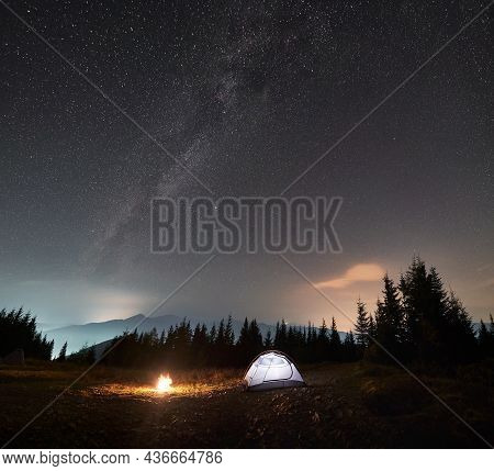 White Shining Tent On Stone Path In The Middle Of Mountain Meadow. Light From Flames Of Bonfire Near