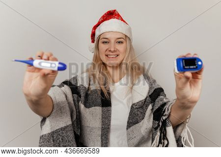 Woman In Santa Hat And Wrap Holds Thermometer And Puls Oximetr On Gray Isolated Background