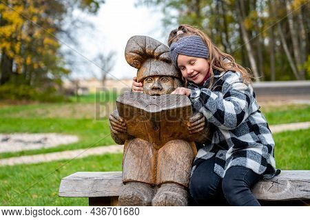 A Girl In The Park On A Bench Next To A Wooden Dwarf Sculpture, The Dwarf Reads A Book, Tervete