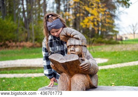 A Girl In The Park On A Bench Next To A Wooden Dwarf Sculpture, The Dwarf Reads A Book, Tervete