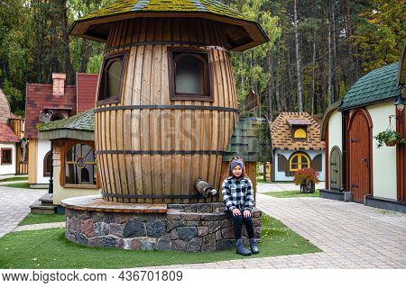 A Girl Sits On A Stone Curb By A Water Tower In A Dwarf Village With Small Colorful Houses, Tervete