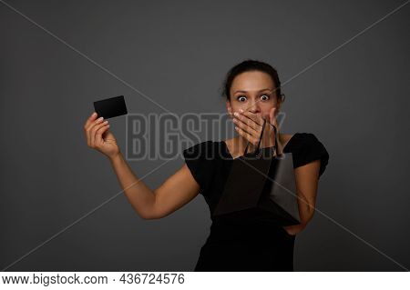 Surprised Woman Holds Black Credit Or Discount Card In One Hand And Covers Her Mouth With Other Hand