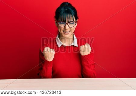 Young brunette woman with bangs wearing glasses sitting on the table excited for success with arms raised and eyes closed celebrating victory smiling. winner concept. 