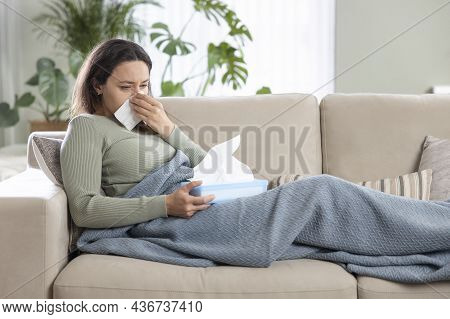 Portrait Of Ill Woman Caught Cold, Feeling Sick And Sneezing In Paper Wipe. She Covered In Blanket W