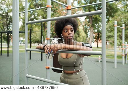 Young restful sportswoman with bottle of water listening to music in earphones on sportsground