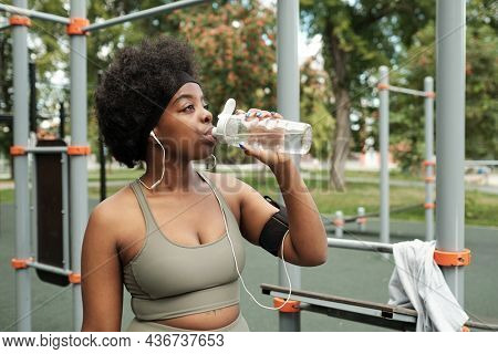 Young thirsty woman in tracksuit drinking water from plastic bottle against sports facilities