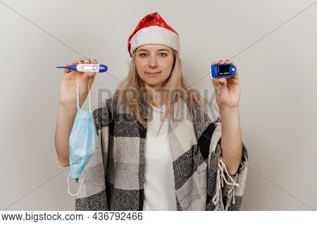 Woman In Santa Hat And Wrap Holds Thermometer And Puls Oximetr On Gray Isolated Background