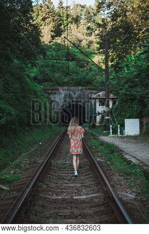 Young Woman In Colourful Dress Walks Along The Rails In The Forest. Lonely Girl Approaches A Tunnel 