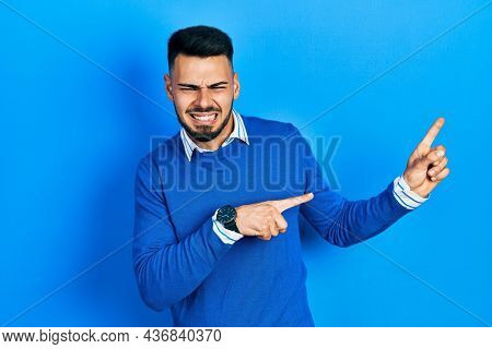 Young hispanic man with beard wearing casual blue sweater pointing aside worried and nervous with both hands, concerned and surprised expression 