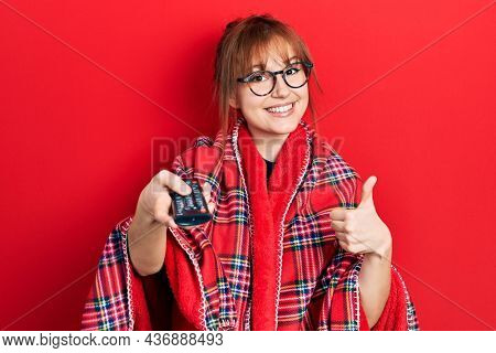 Redhead young woman wrapped in a red warm red blanket using tv control smiling happy and positive, thumb up doing excellent and approval sign 