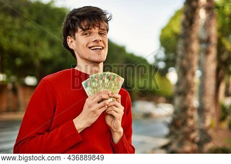 Young hispanic man smiling happy holding argentinian pesos banknotes at the city