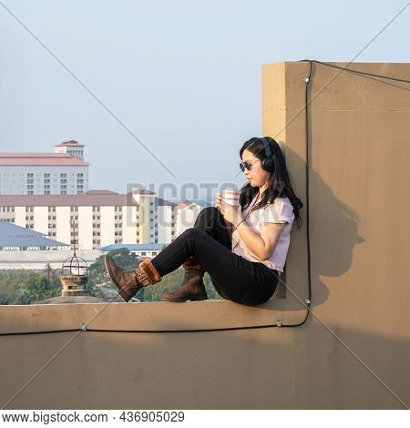 Glasses Asian Woman Is Listening Music Via Headset, Drinking Coffee On Rooftop Barricade Cement (not
