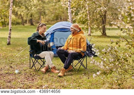 camping, tourism and travel concept - happy couple with thermos pouring tea to cup at tent camp