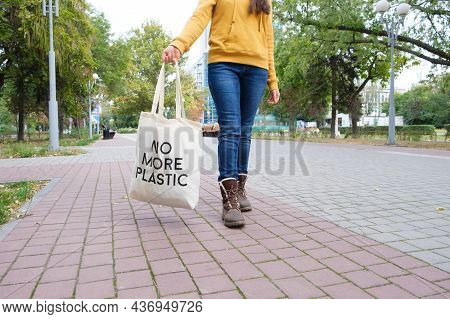 A Woman Walks Down The Street And Holds A Reusable Cloth Bag That Says No More Plastic.