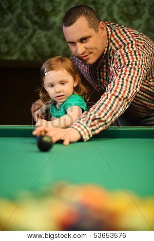 A man in checkered shirt teaches his daughter to play billiards