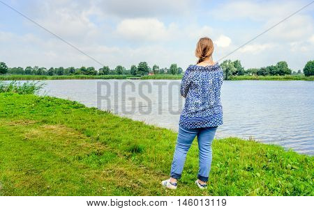 Woman with a long hair braid and decorated a blue shirt stares over the water of a small lake. It's a sunny day in the summer season.