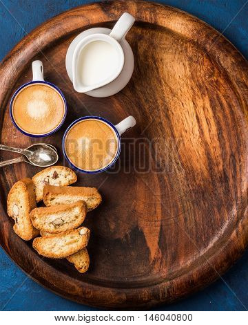Coffee espresso in cups with cookies and milk in jug on wooden serving round board over dark blue painted plywood background, top view, copy space