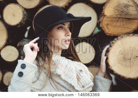 Gorgeous young blue eyed female model in autumn. Beautiful young woman in black floppy hat, beige sweater and coat posing next to tree chunks. Matte filter, neutral colors.