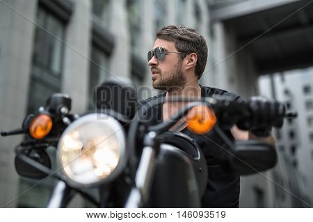 Charismatic man sits on the black motorbike on the skyscraper background. Headlamp switched on. He wears a black T-shirt, black gloves and sunglasses. He looks to the right with parted lips. He holds hands on the rudder. 
