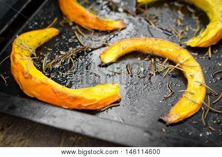 Freshly baked and juicy slices of pumpkin with addition aromatic herbs and olive oil on a baking tray. Pumpkin slices arranged on rows. Thanksgiving Halloween holidays concept. Selective focus.