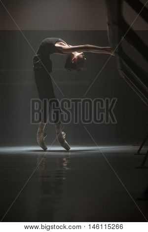 Young ballerina in a dance hall. She is in ballet shoes and black wear. She leans on the barre while makes a pose in front of the wall.Light falls on her neck and chest. Low key photo.