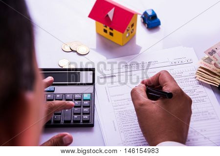 Indian man or accountant person filing Indian income tax returns form or ITR document showing indian currency, house model, toy car and calculator over white table top, selective focus