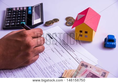 Indian man or accountant person filing Indian income tax returns form or ITR document showing indian currency, house model, toy car and calculator over white table top, selective focus
