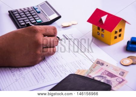 Indian man or accountant person filing Indian income tax returns form or ITR document showing indian currency, house model, toy car and calculator over white table top, selective focus