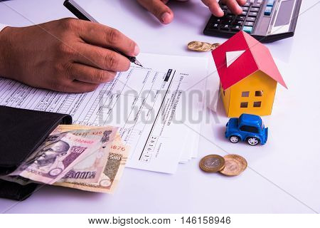 Indian man or accountant person filing Indian income tax returns form or ITR document showing indian currency, house model, toy car and calculator over white table top, selective focus