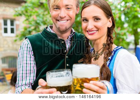 Couple, women and man, wearing Bavarian clothes is clinking beer glasses in a beer garden 