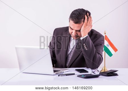 stressed indian businessman holding head with one hand while filling tax form or accounting on laptop