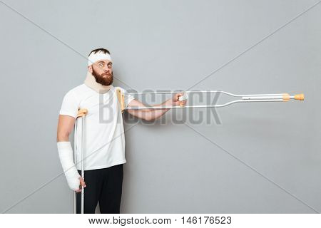 Funny bearded young man standing and pointing away with crutches over white background