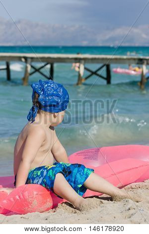 Adorable little kid boy sitting on a pink mattress for swimming and looking at the sea and wooden pier. Summer vacation at the ocean. Child sitting on the sand beach and enjoying outdoor swimming.