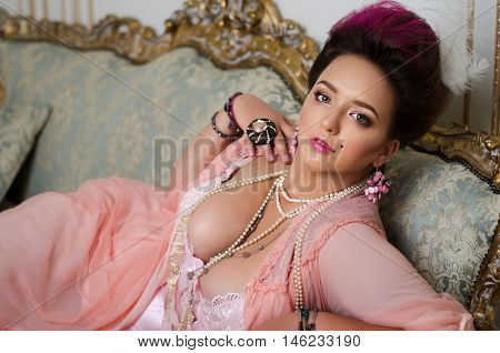 beautiful brunette with curvaceous Rococo hair rings and beads lies on a beautiful sofa in pink peignoir.