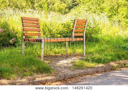 Horizontal photo of interesting bench from metal and wood which is placed on the lawn next to pavement (sidewalk). Bench has two backrests which allows to sit face to face.