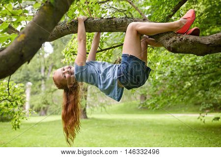 friendship, childhood, leisure and people concept - happy smiling little redhead girl hanging upside down on tree in summer park