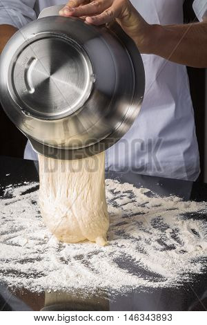 Chef pouring out freshly beaten dough mixture from a stainless steel mixing bowl onto a floured kitchen counter while making fresh bread