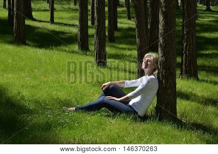 Woman resting on spring grass leaning against a tree in coniferous forest and absorbing spring sunbeams
