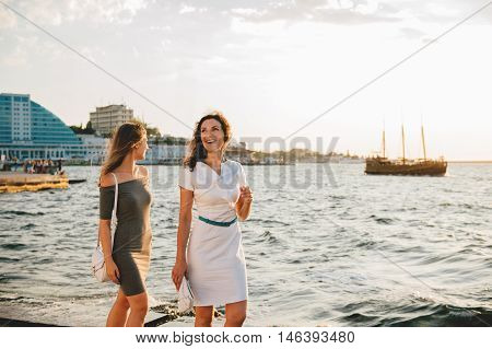 Mother and teen daughter walking talking and smiling on the seafront of city. Sunset light.