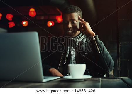 Indoor portrait of young black man sitting in cafe, drinking coffee or tea and working with lap top. Model looking at screen, expressing joyful and excitement with face. Toned.