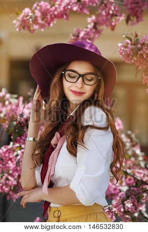 Outdoor portrait of young beautiful happy smiling lady posing near flowering tree. Model wearing stylish accessories and clothes. Girl closed her eyes. Female beauty and fashion concept. City lifestyle.