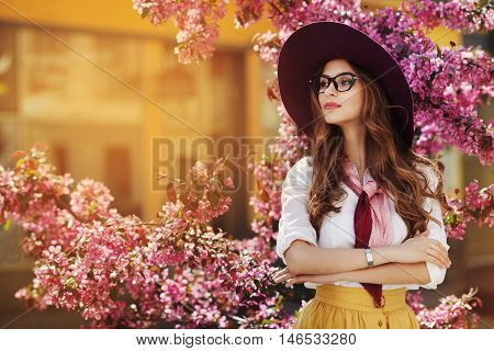 Outdoor portrait of young beautiful fashionable lady posing near flowering tree. Model wearing stylish accessories and clothes. Girl looking aside. Female beauty, fashion concept. City lifestyle. Copy space for text.