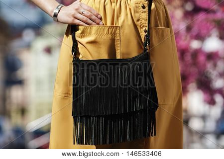 Elegant outfit. Closeup of the small black suede bag with fringe. Boho style. Fashionable girl on the street. Female fashion. City lifestyle.