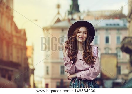 Outdoor portrait of young beautiful fashionable smiling lady posing on old street. Model wearing stylish hat and clothes. Sunny day. Female fashion. City lifestyle. Toned style instagram filters.