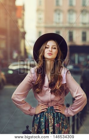 Outdoor portrait of young beautiful fashionable playful lady posing on old street. Model wearing stylish hat and clothes. Sunny day. Female fashion. City lifestyle. Toned style instagram filters.