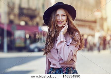 Outdoor portrait of young beautiful fashionable happy lady posing on a street of the old city. Model wearing stylish wide-brimmed hat and clothes. Girl looking at camera. Female fashion. City lifestyle.