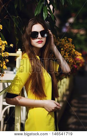 Outdoor portrait of young beautiful fashionable lady posing on street. Model wearing sunglasses and stylish yellow summer dress. Girl looking at camera. Female fashion concept. City lifestyle. Sunny day.