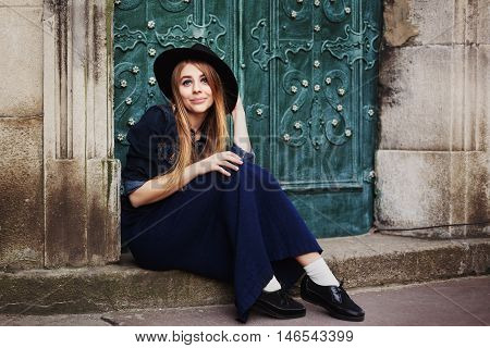 Street full body portait of stilylish smiling young woman sitting near the door. Model looking at camera. City lifestile.