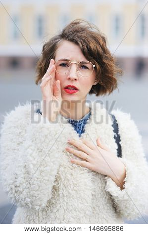 Young beautiful short-sighted girl in a white jacket stares through his glasses at a city street, close-up.